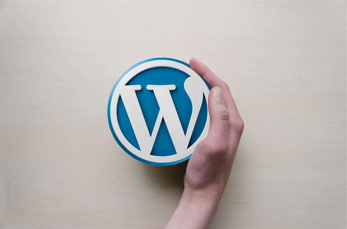 A hand holding a round blue WordPress logo badge against a pale wooden background.