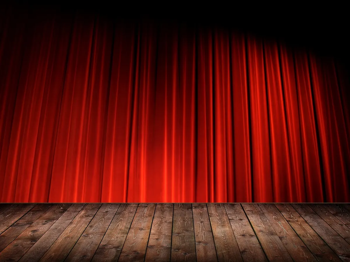 A closed red theatre curtain above an empty wooden stage, the space where a show is prepared before the audience sees it.