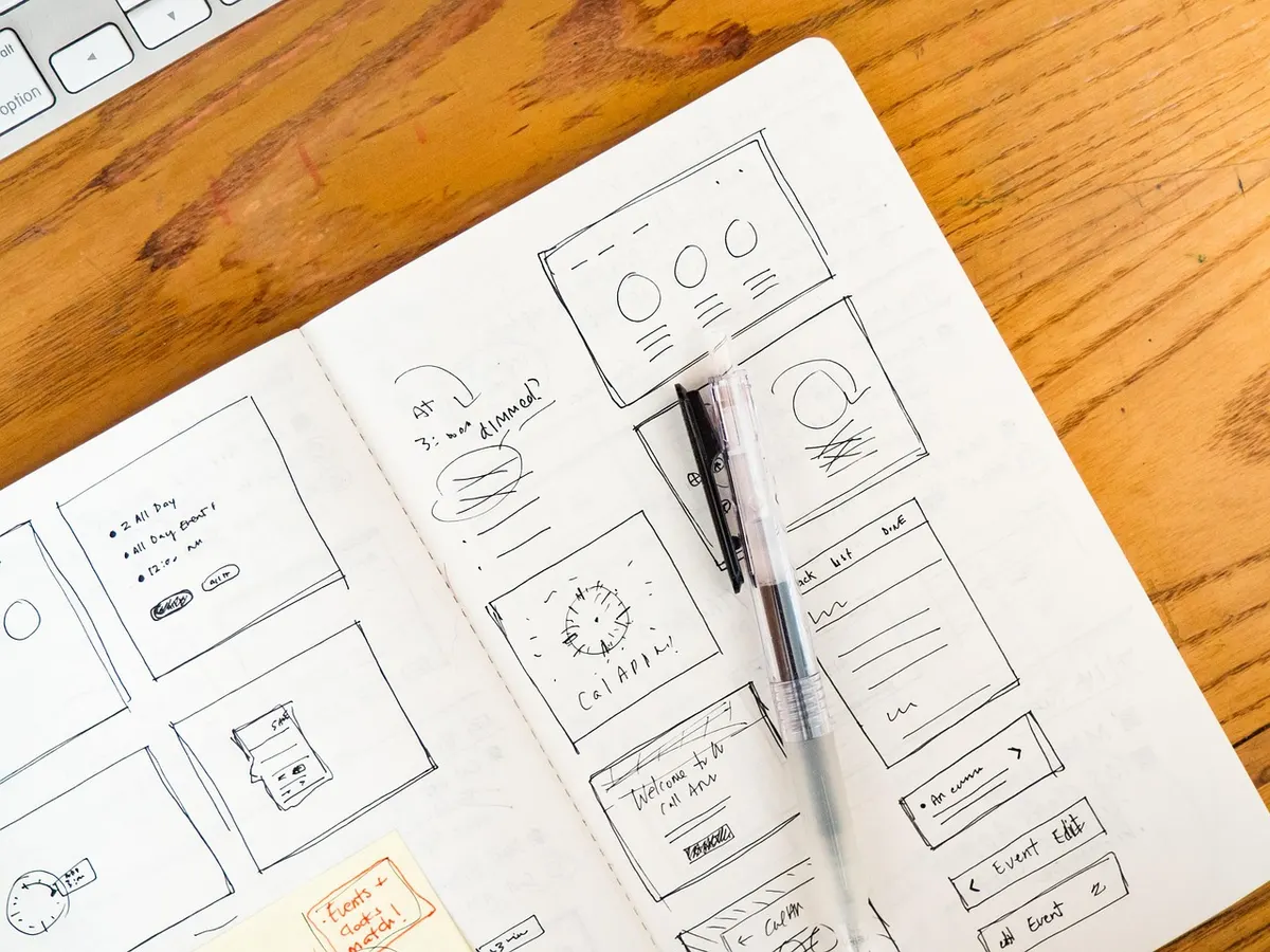 Hand-drawn website wireframes in an open notebook on a wooden desk, the kind of rough layout sketch UX design starts with.