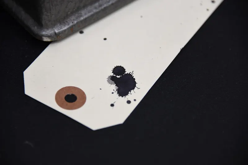 A blank white price tag with a metal eyelet and a few ink spots, lying on a dark surface.
