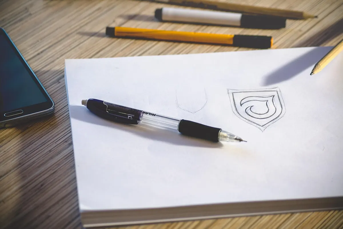 A rough logo sketch of a shield shape on a sketchpad with a pen beside it, the early stage where a designer explores ideas before anything goes digital.