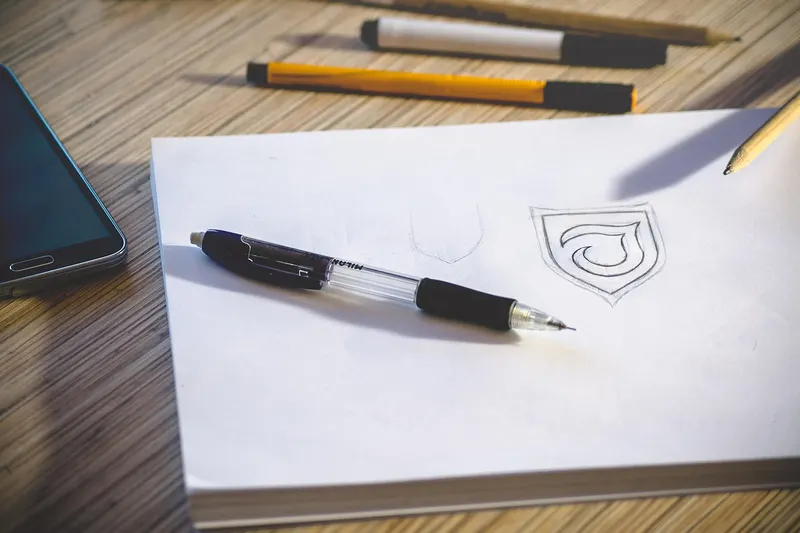 A rough logo sketch of a shield shape on a sketchpad with a pen beside it, the early stage where a designer explores ideas before anything goes digital.