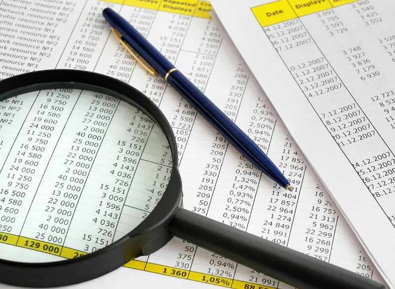 Magnifying glass and pen resting on printed spreadsheets, ready to check the details